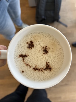 Hand, die eine Tasse Kaffee hält, mit einem lächelnden Gesicht, das kreativ mit Kaffeesatz auf dem Schaum gezeichnet ist – ein Moment der Entspannung in einer Online-Agentur.
